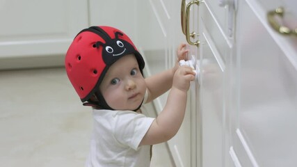 Childproofing a house, baby wearing safety helmet exploring kitchen cupboards, curious toddler trying to open a cabinet door with installed child safety lock. High quality 4k footage