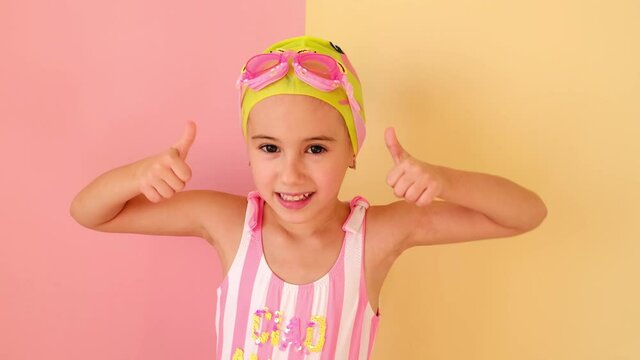A child in a swimming cap and swimming glasses shows a class, demonstrating success gesture, thumb up with hand. Cute Caucasian girl in pink swimsuit. Children's swimming lessons. Playing sports