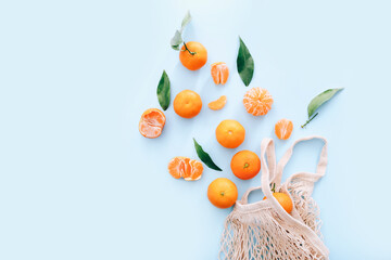 Fresh clementines with leaves scattered from string bag on blue background, top view