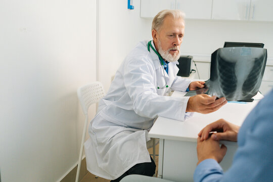 View from back of unrecognizable male patient listening mature adult doctor with chest x-ray lungs scan giving professional medical consultation for treatment illness, explaining causes of disease.