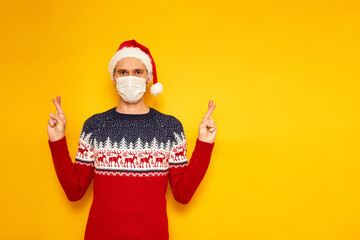 Fototapeta na wymiar man in New Year sweater, red Christmas hat, medical mask stands isolated on yellow background with space for text with his fingers crossed and making wish. concept - holiday, pandemic, social distance