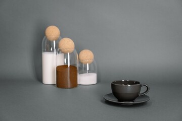 Fototapeta na wymiar Coffee cup and ingredients in glass jars on grey background