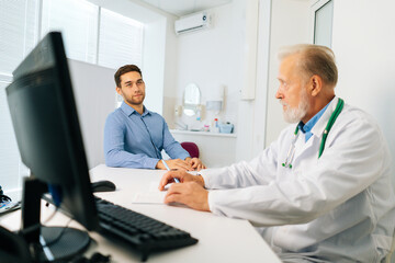 Serious mature adult male doctor in medical coat consulting young man patient about illness or surgery sitting at table in medical office with light interior, during checkup visit.