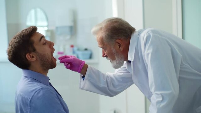 Mature male doctor testing sick young man patient for for possible coronavirus infection using throat swab during checkup visit in clinic office. Concept of Covid diagnostic. Shooting in slow motion.