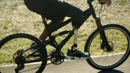 Side view of unrecognisable man with bionic leg cycling in park. Tracking shot of young man riding bicycle along path in public park, spending weekends outdoor. Disability, challenge, sport concept - Powered by Adobe