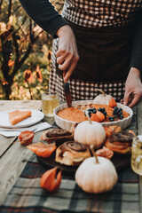 The woman cuts and serves the pie to guests. Thanksgiving festive table. Cozy autumn style table setting with pumpkins and leaves. Pumpkin pie and cinnamon rolls. Fall styled composition.