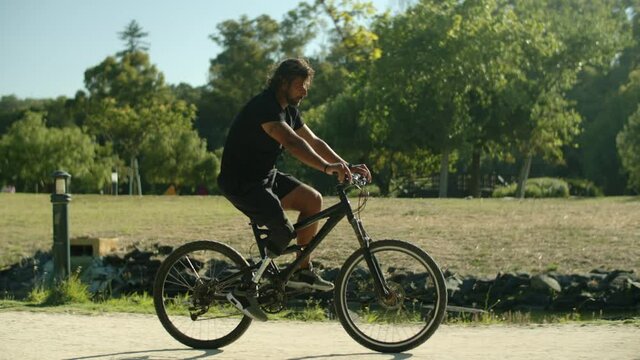 Side view of young Caucasian man with disability cycling in park. Tracking shot of adult with bionic leg riding bicycle, taking care of health by doing physical activity. Disability, sport concept 