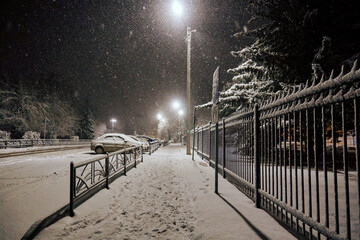 Winter, white fluffy snow powdered all around, falling snow. Night, lights shine along the black metal fence. Soft selective selective focus
