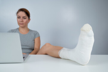 Caucasian woman lifted her leg with plaster to work desk and works on laptop on white background