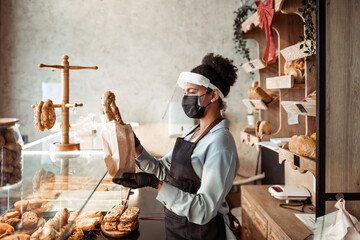 African American middle aged female worker with shield and protective mask on face working in bakery. Coronavirus, Covid-19 concept.