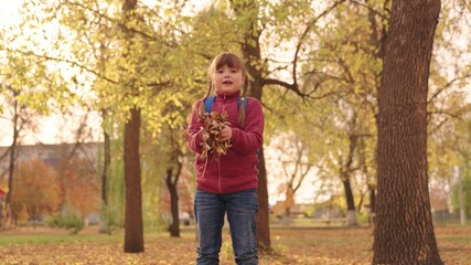 little happy girl with a backpack throws leaves high up and laughs cheerfully, child play in the autumn park, kid walks in the city park, raise her face looking at the golden leaf fall, autumn season