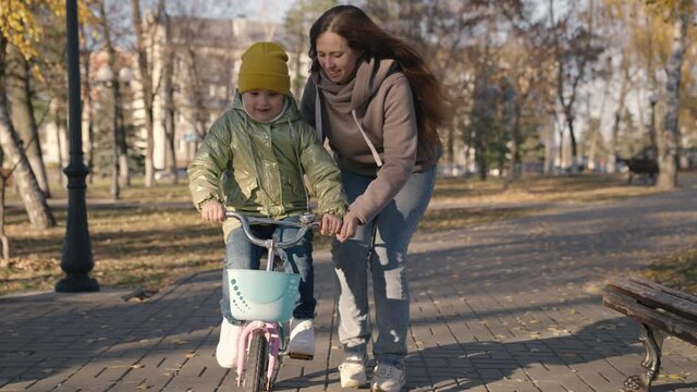 mother teaches little child to ride bike in the city, kid rides on sidewalk, happy family, baby plays and laughs with mom, learn ride, make childhood dream come true girls, daughter and mom are funny