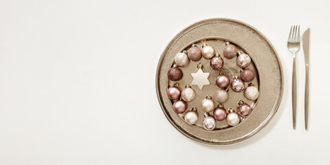 Christmas table decorations with shining small balls beige pink color in metal plate, fork, knife on white background, wide banner with copy space. New Year winter holiday food concept