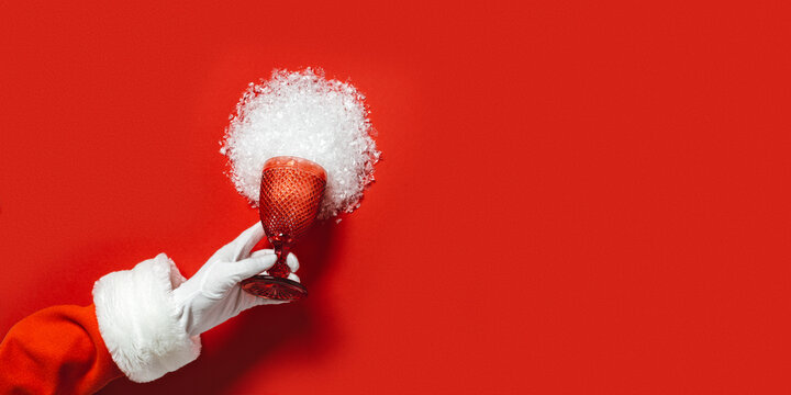 Santa hand hold glass from red glass from which snow scattered, creative concept of winter holidays, New year, Christmas party. Banner with colour glassware