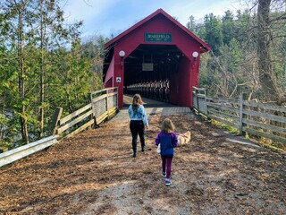 Fototapeta premium A mother and daughter walking together through the red Wakefield covered bridge along the Gatineau River on a beautiful sunny fall day in pretty town of Wakefield, Quebec, Canada.