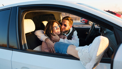 Loving carefree tourists travelers couple sitting in car at sunset in countryside. Man hugs and kisses his girlfriend while stopping on trip. Travel, road trip and comfortable concept
