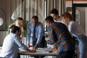 Motivated concentrated young mixed race teammates gathered at table, discussing sales data statistics or marketing research results, analyzing paper reports or documents together in modern boardroom.