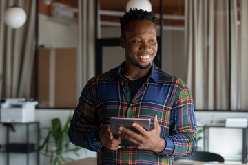 Joyful young dreamy african american biracial male employee worker holding digital computer tablet in hands, reading media news, using corporate software applications. modern tech usage concept.
