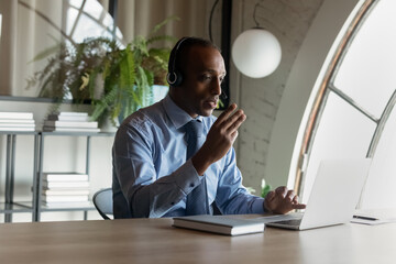 Concentrated young african american businessman coach in headset mentoring colleagues online, holding distant video call meeting or sharing knowledge distantly using computer app in modern boardroom.