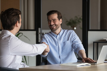 Happy two diverse colleagues shaking hands, celebrating making agreement at office meeting. Smiling businessmen establishing partnership at workplace, successful cooperation collaboration concept.