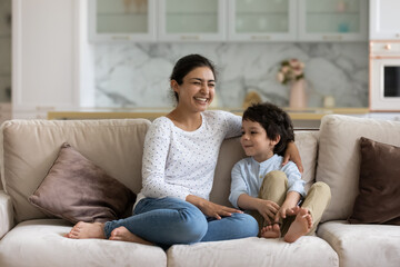 Obraz premium Laughing Indian mom enjoying leisure time with happy son at home, talking to boy. Mother and cheerful preschool child having fun together, relaxing on couch. Babysitter watching cute kid