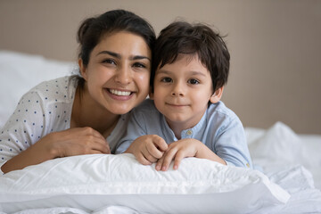 Obraz na płótnie Canvas Happy Indian mom and kid lying on bed, resting close in white linen, looking at camera, smiling. Younger mother and little preschool son having fun, enjoying leisure in bedroom. Home portrait
