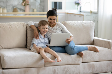 Happy Indian babysitter watching kid, showing online movie on laptop to child. Cheerful mom and adopted little son resting on sofa at home, making video call on computer, shopping on internet