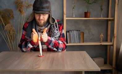 A Caucasian bearded man dressed in a warm winter hat and a scarf sits at home at a table and warms his hands from a burning candle.