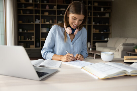 Happy beautiful young woman writing notes in copybook, improving knowledge studying distantly on online courses, using computer and books, preparing for examination, domestic education concept.