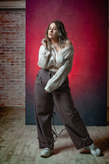 Super sexy, happy, fashionable and stylish girl plus size poses in a satin blouse with a deep neckline and bare shoulder, gray trousers and jewelry accessories on a red-pink gradient background 