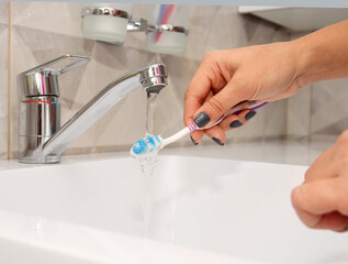 A woman holds a toothbrush and washes it under water in the bathroom sink under the faucet.