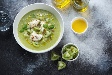 Bowl of green tomato gazpacho with mozzarella cheese, above view on a dark-grey stone background...