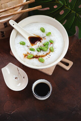 Beige bowl with asian congee or conjee porridge, edamame and soy sauce, high angle view on a rusty metal background, vertical shot