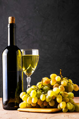 a glass glass and a bottle of white wine and a brush of white grapes on a cutting board on a black background