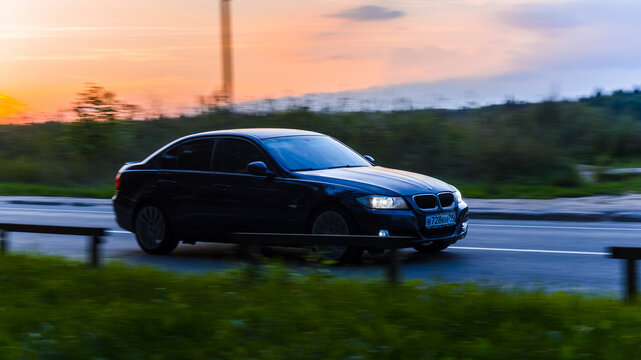 BMW 3 series E90 driving on the country road at dusk. Front side view of black BMW 330i in motion on the background of evening sunset sky