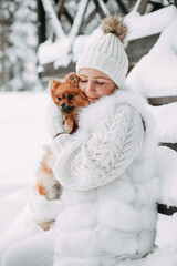 In the winter forest, a young blonde girl with long hair in a white sweater, white fur coat, white hat and white mittens smiles and holds a Spitz dog in her arms
