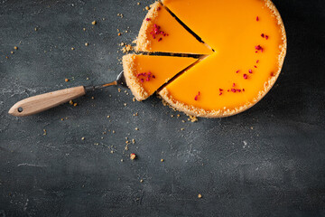 Flat lay of cheesecake with yellow mango flavoured layer decorated with red crumbs of freeze-dried raspberries. Cake cut on the dark marble background