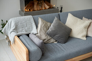 Various cushions and plaid on gray sofa with fireplace on the background. Home interior, cozy living room, room decor, scandinavian style concept.