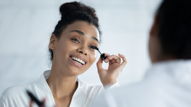 Smiling beautiful young african american woman looking in mirror applying black brown mascara on lashes in morning after shower, getting ready doing daily nude makeup routine in bathroom at home. - Powered by Adobe