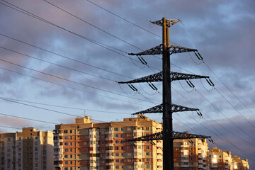 High voltage tower with electrical wires on background of residential buildings and evening sky. Electricity transmission lines in a city, power supply concept