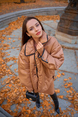 Fashion full length portrait from upper point of view of elegant fashionable woman in beige coat standing in fountain with yellow autumn foliage