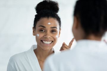 Mirror reflection attractive young african american woman in bathrobe applying moisturizing smoothing cream on face, excited by effective cosmetics results, enjoying morning skincare beauty routine. - Powered by Adobe