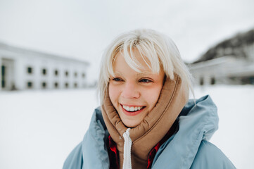 Portrait of a positive blonde girl with white eyebrows in warm clothes on a winter street, looking away and smiling.