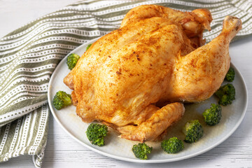 baked whole chicken with broccoli on a round platter on a white wood background