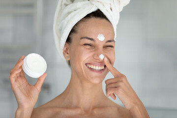 Head shot joyful young hispanic woman applying moisturizing cream on face, doing morning skincare...