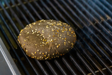 Brown bread for hamburger with sesame seeds is fried on the grill. Thin smoke coming from under the bun. Close-up.