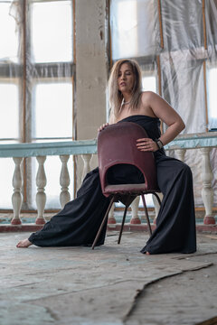 Super stylish beautiful curvy girl plus size in an evening jumpsuit dress in an abandoned old theater with columns posing sitting on a chair barefoot