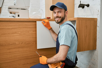 Cheerful male worker fixing cupboard door in kitchen