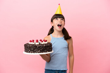 Little caucasian kid holding birthday cake isolated in pink background looking up and with...