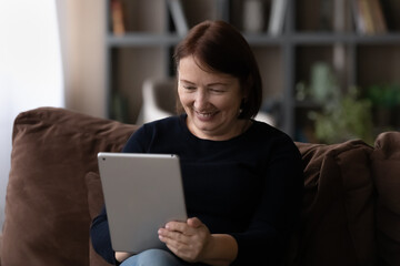 Close up smiling mature woman using tablet, sitting on couch, happy senior female looking at modern device screen, chatting in social network, watching video, enjoying leisure time with gadget